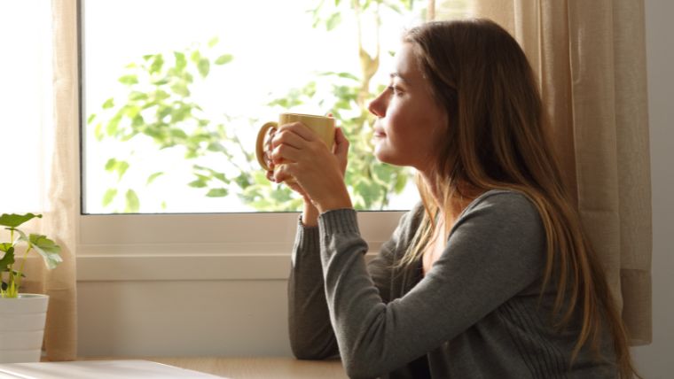 Woman drinking a warm beverage while looking out the window on a sunnday day | TMS Side Effects Important Facts You Should Know | Mindful Health Solutions