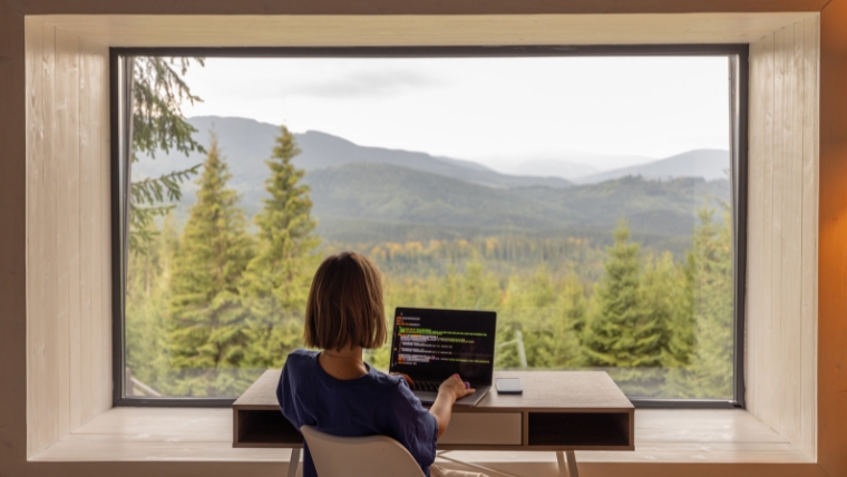 Woman works on laptop while sitting by the table in front of panoramic window with great view on mountains | How to Effectively Juggle Work and Well-Being While Managing Depression | Mindful Health Solutions