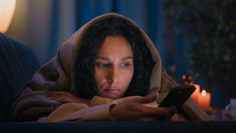 The-Psychology-of-Comparison-Why-We-Do-It-and-How-to-Stop Woman of color on couch under blanket scrolling phone with a sad expression | The Psychology of Comparison Why We Do It and How to Stop | Mindful Health Solutions