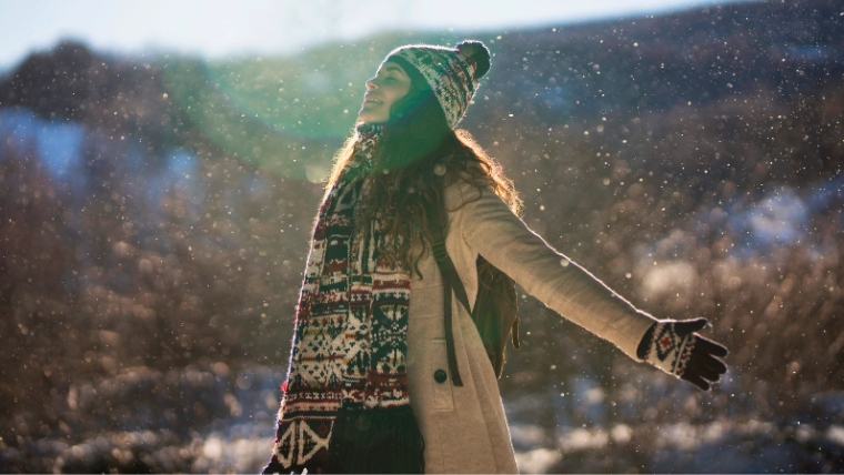 Woman standing outside in woods while it is snowing. Her arms are out and her head is titled up, enjoying the snow fall. | Boost Your Well-being with 9 Mindfulness Practices This Holiday Season | Mindful Health Solutions